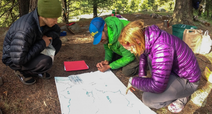 Three people examine a map that is spread out on the ground in a wooded area. 